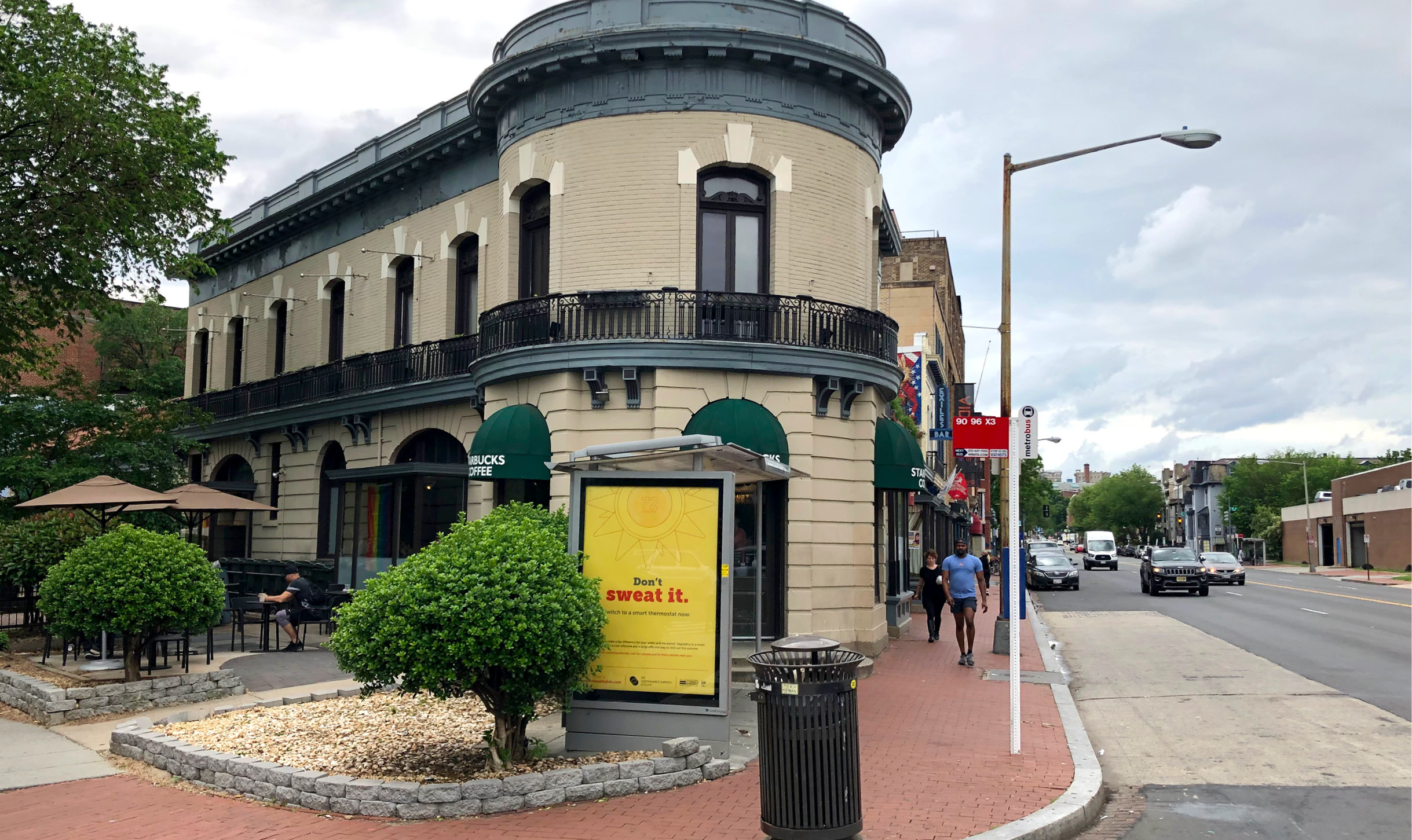 A wide shot of a bus stop in front of an old brick building with a DCSEU ad in it reading Don't Sweat It that has a sun with a smart thermostat center at the top. In the background there are people walking down the street.